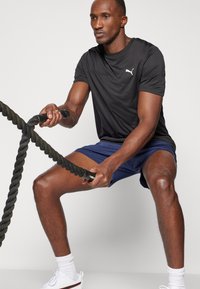 Black athletic T-shirt with white logo, paired with navy shorts. Individual using thick black exercise ropes, displaying muscular arms.