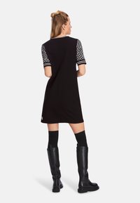 Schwarzes Kleid mit kurzen gemusterten Ärmeln, mit geradem Schnitt. Kombiniert mit knielangen schwarzen Stiefeln und knielangen Socken.