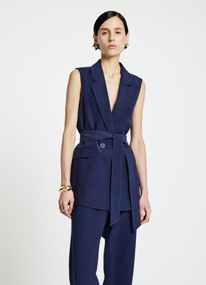 Blazer smanicato blu navy con profondo scollo a V, vita con cintura e tasche laterali. Vestibilità sartoriale, realizzato in tessuto strutturato, finitura liscia.