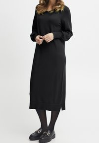 Svart långärmad stickad klänning med rund halsringning, avslappnad passform och sidslitsar. Kombinerad med svarta plattform-loafers och strumpbyxor.