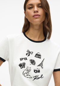 T-shirt de algodão branca com gola preta em ringer. Apresenta estampas em estilo de esboço, em preto, de um gato, uma câmera, a Torre Eiffel e diversos acessórios.