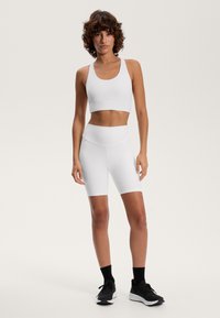 Biele športové podprsenky a vysoko pásové šortky vyrobené z elastického materiálu. Model má na sebe čierne tenisky, ktoré zdôrazňujú štýlový a priliehavý dizajn.