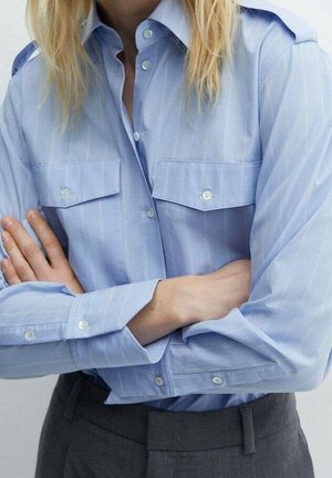 Osoba z założonymi skrzyżowanymi rękami, ubrana w jasnoniebieską koszulę na guziki z kieszeniami na piersi oraz ciemnoszare spodnie.