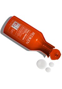 Shampoo Redken Frizz Dismiss in una bottiglia di plastica arancione con tappo a scatto, tre ciuffetti circolari bianchi di shampoo accanto ad essa.