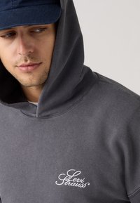 Γκρι φούτερ με κουκούλα και ριμπ τελείωμα στο λαιμό, με λευκό κεντητό λογότυπο στο στήθος. Λείο ύφασμα. Φοράει σκούρο καπέλο.