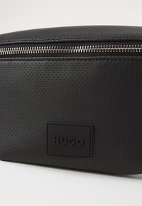 Fekete perforált anyagú szükségleteket tartó táska ezüst cipzárral és fekete, „HUGO.” felirattal domborított téglalap alakú címkével. Lekerekített tető kialakítás.