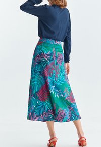 Gonna maxi floreale e colorata con sfondo blu, caratterizzata da motivi di piante verdi e rosa. Indossata con una maglia a maniche lunghe scura e sandali rossi.
