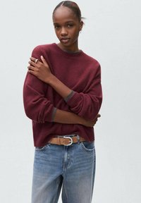 Burgundska pletena jopica z okroglim izrezom in rebrastimi manšeti, v kombinaciji s svetlo moderimi kavbojkami in svetlim pasom z srebrno sponko.