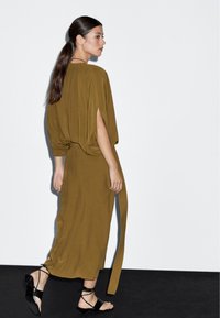 Olivově zelené, volně střižené šaty s krátkými rukávy ve stylu netopýřích křídel, s vypasovaným pasem a zavázaným detailem na zádech, doplněné černými sandály na řemínky.