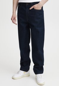 Temno modri denim kavbojke z ohlapnim krojem, rumenim šivom, sprednjim žepom in kombinirane z belimi supergami.