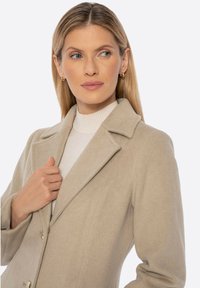 Beżowy płaszcz z mieszanki wełny z wyciętym kołnierzem, frontowymi guzikami i bocznymi kieszeniami. Pod spodem biały, prążkowany golf. Gładka faktura.