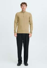 Maglione beige in lana con colletto alto e dettaglio a mezza zip, abbinato a pantaloni neri a gamba larga e scarpe nere. Texture liscia, stile aderente.