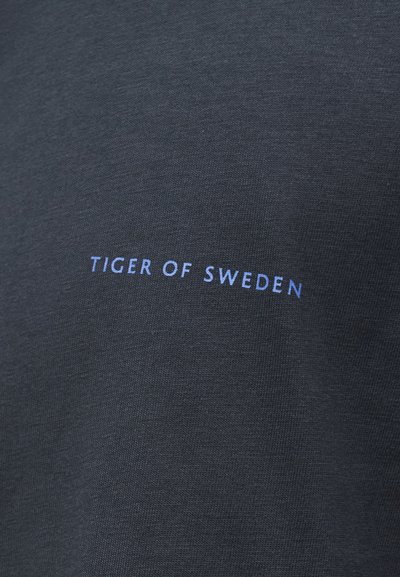 Σκούρο γκρι ύφασμα με μικρό ανοιχτό μπλε κείμενο που αναγράφει "Tiger of Sweden" στο κέντρο της επιφάνειας.