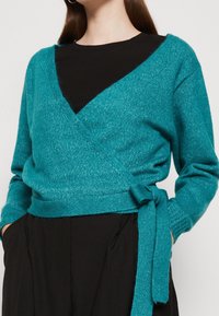 Teal insluten tröja i mjukt material, med V-ringning, långa ärmar och en knut i midjan som framhäver formen.