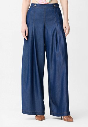 Vrouw draagt donkerblauwe denimbroek met hoge taille, wijde pijpen, zijknopen en plooien, gecombineerd met geweven puntige hakken.