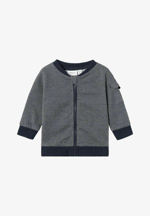 Felpa grigia con zip in tessuto texturizzato, polsini e colletto a coste in navy, e una piccola tasca sul maniche sinistro. Design semplice e casual.