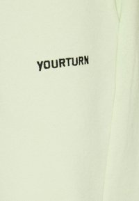 Světle zelená tkanina s hladkým povrchem má na jedné straně černý vyšitý text "YOURTURN".