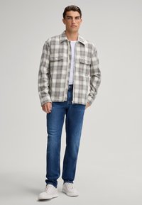 Plaid flannel shirt with button-down collar, gray and beige tones, two front pockets, worn over a white t-shirt, paired with blue jeans and white sneakers.