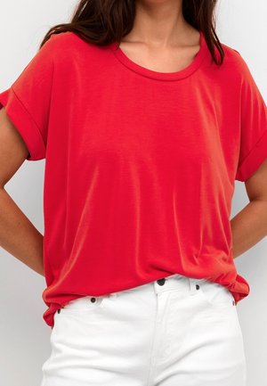 Vrouw draagt een felrode korte mouw t-shirt dat lichtjes in een high-waisted witte broek is gestopt, staand met handen in de zij.