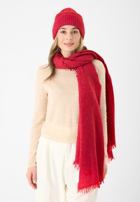 Gehaakte rode muts en sjaal, beide met franjes aan de uiteinden. De sjaal heeft een kleine hartaccent. Een beige trui en witte broek maken de outfit compleet.