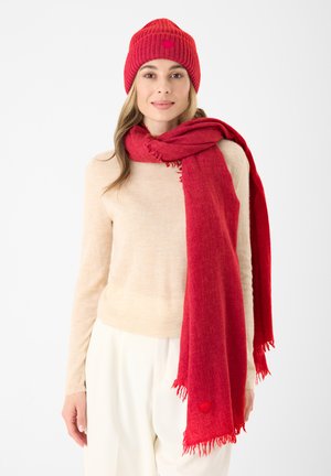 Bonnet en tricot rouge et écharpe, tous deux avec des extrémités à franges. L'écharpe présente un petit accent en forme de cœur. Un pull beige et un pantalon blanc complètent la tenue.