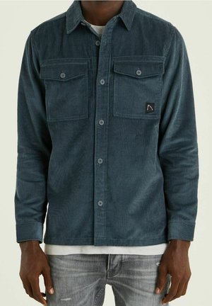 Mand iført en mørkeblå kordfløjls skjorte med knapper og to brystlommer samt lysegrå slidte jeans.