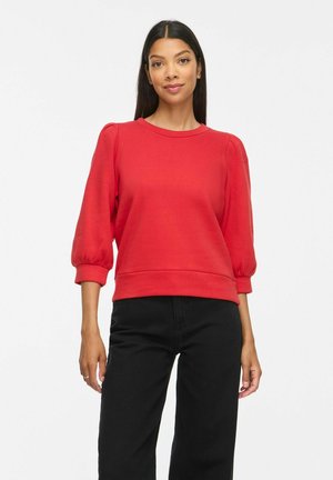 Femme aux longs cheveux foncés portant un haut rouge à manches bouffantes et un pantalon noir, debout devant un fond blanc uni.