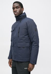 Granatowa ocieplana kurtka z wysokim kołnierzem, licznymi kieszeniami i gładką teksturą. Wykończona czarnymi okuciami i guzikami.