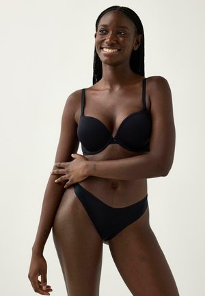 Kvinde med flettet hår iført sort bh og undertøj, smilende, stående mod en ensfarvet lys baggrund med den ene arm krydset over kroppen.