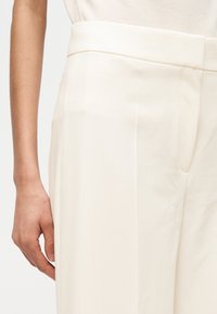 Λεπτός δεξιός βραχίονας δίπλα σε κρεμ ψηλόμεσο παντελόνι με πιέτα μπροστά και ζώνη μέσης.