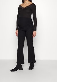 Czarny prążkowany top z długim rękawem z koronkowym wykończeniem oraz czarne spodnie w stylu dzwonów. Modelka ma na sobie czarne botki, prezentując dopasowany styl w ciąży.