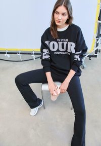 Čierna mikina s šedým textom "JSEM TVOJÍ INŠPIRÁCIOU," skombinovaná s čiernymi nohavicami a bielymi teniskami. Sedí na bielej stoličke.