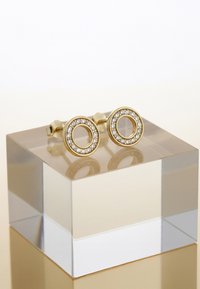 Guld cirkulära studörhängen med en ihålig mitt, prydda med små klara ädelstenar runt den yttre kanten, visade på en klar akrylblock.