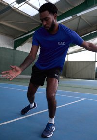 Camiseta atlética azul com o logótipo EA7, calções pretos e ténis azuis. O estilo é ajustado e foi concebido para movimentos num campo de ténis.