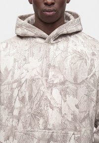 Gros plan d'une personne portant un sweat à capuche beige avec un motif camouflage léger de feuilles et de branches, et une poche kangourou à l'avant.