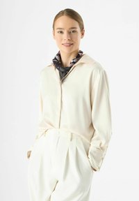 Cremefarbene Seidenbluse mit Kragen; verfügt über lange Ärmel mit geknöpften Manschetten und einem schwarzen gemusterten Schal um den Hals. Weiße, hoch taillierte Hose.