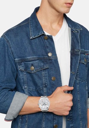 Orologio cronografo argentato con quadrante bianco, bracciale in metallo e dettagli neri, indossato su un polso. Giacca in denim blu con bottoni in bronzo visibili.