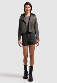 Sieviete, tērpusies tumši pelēkā ādas žaketē pār pelēku topu, melnās džinsa šortos un melnās mežģīnēs iesietās kaujas zābakos, stāvot pret balto fonu.