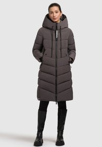 Grå pufferjakke med chevron-quiltet design, en lang lynlås, hætte med snoretræk og sidelommer. Kåret med sorte knæhøje støvler.