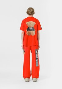 Camisa roja de manga corta con un gráfico de oso de peluche en la parte posterior, combinada con pantalones de corte holgado a juego. Incluye detalles de tela únicos y etiquetas.