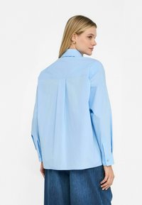Lichtblauw shirt met een ontspannen pasvorm, lange mouwen en knoopmanchetten. Bevat een plooi-detail op de achterkant. De stof lijkt glad en fris.
