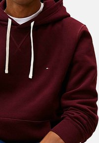 Felpa color marsala con tasca frontale, cordini bianchi e un piccolo dettaglio del logo. Tessuto morbido con polsini e orlo a coste.