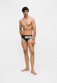 Jeune homme debout, pieds nus, portant un slip noir avec un grand texte blanc "DSQU" sur la ceinture, sur un fond blanc uni.