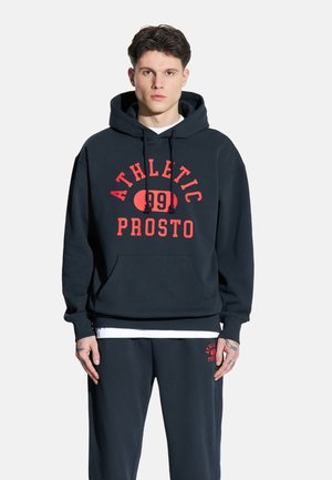 Granatowy hoodie z kapturem na sznurki i przednią kieszenią. Na przodzie czerwony napis "ATHLETIC 99 PROSTO." Noszony z pasującymi granatowymi dresami.