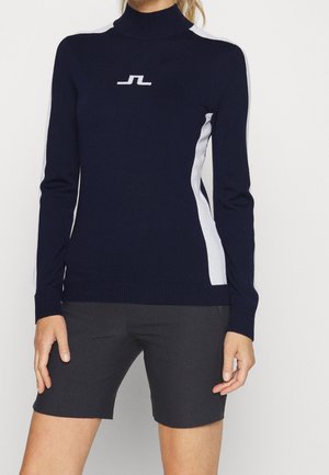 Kvinde iført en mørkeblå langærmet sweater med hvide sidestriber og logo, kombineret med mørkegrå tætsiddende shorts.