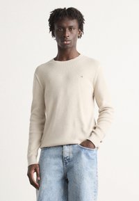 Beige ribbed knit sweater with a round neckline, long sleeves, and a small logo accent on the left chest. Worn with light blue denim jeans.