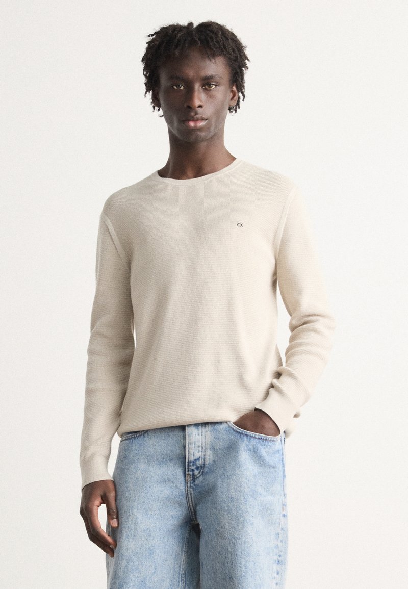Beige ribbed knit sweater with a round neckline, long sleeves, and a small logo accent on the left chest. Worn with light blue denim jeans.