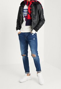 Chaqueta bomber de cuero negra, sudadera blanca, jeans rasgados azules, bufanda a rayas rojas y marinas, y zapatillas blancas. Look casual con texturas en capas.