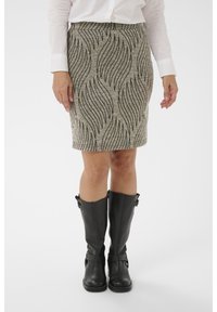 Femme portant une jupe beige avec un motif de lignes ondulées noires, associée à des bottes noires montantes et une chemise blanche à manches longues.