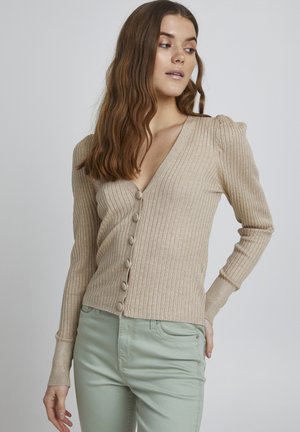 Beige Rippstrickjacke mit V-Ausschnitt, sechs vorderen Knöpfen und gepufften Schulterakzenten, kombiniert mit hellgrünen Hosen.